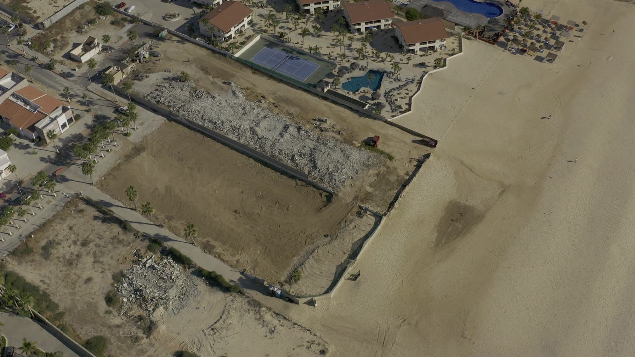 Overhead drone frame in Baja California Sur, Mexico showing sandy parcels, red-roofed buildings, pools, a road, and no visible people.