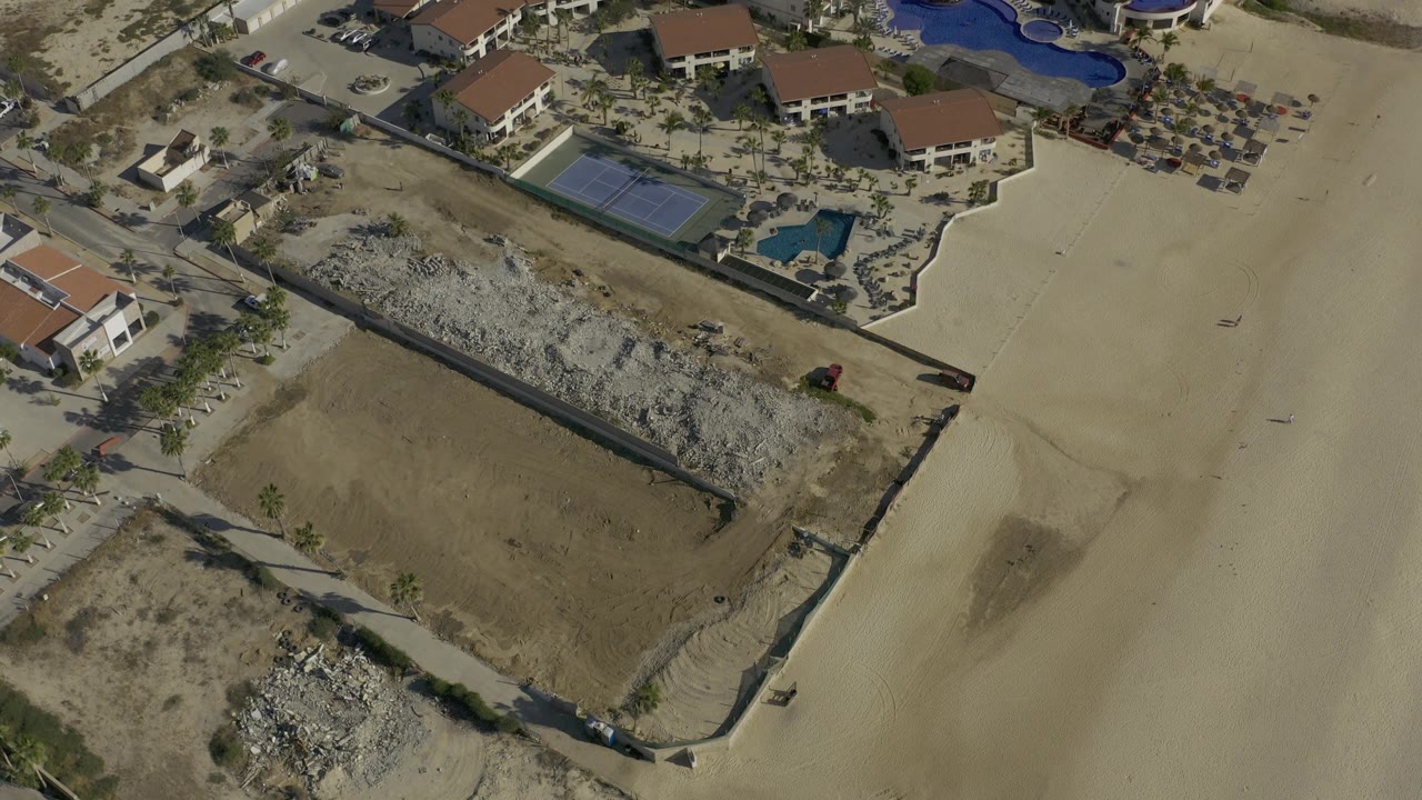 High aerial frame in Baja California Sur, Mexico with open sand in the foreground, residential buildings and pools above it, and no visible people.