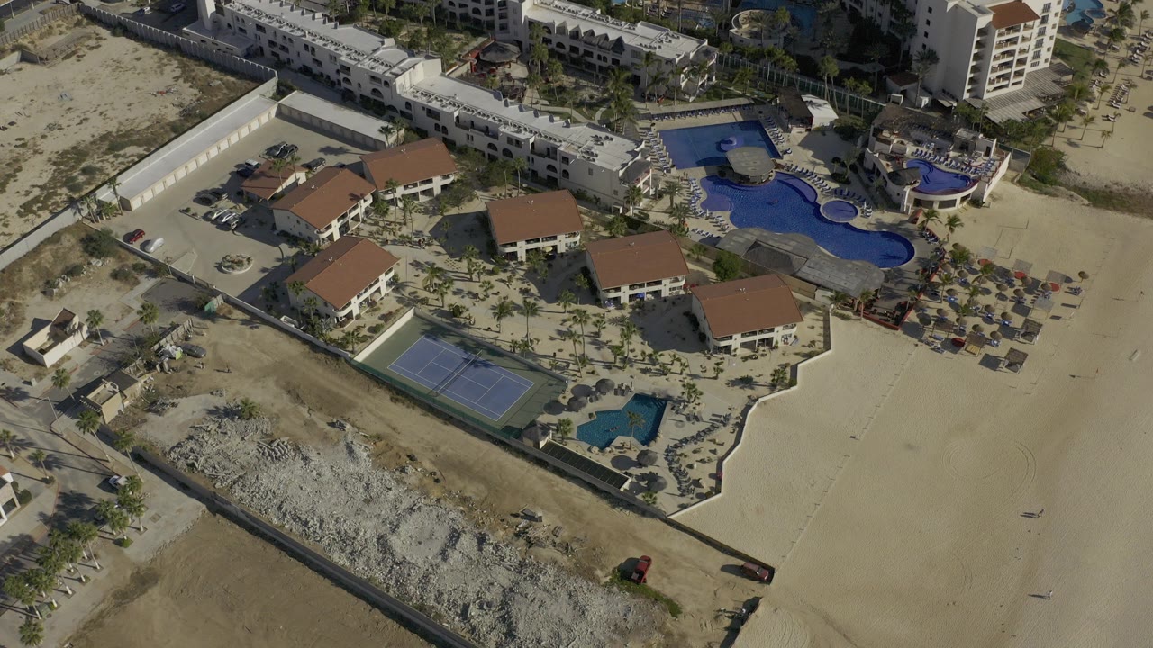 High aerial frame over Baja California Sur, Mexico showing white resort-style buildings, red roofs, blue pools, a tennis court, sandy open ground, and no visible people.