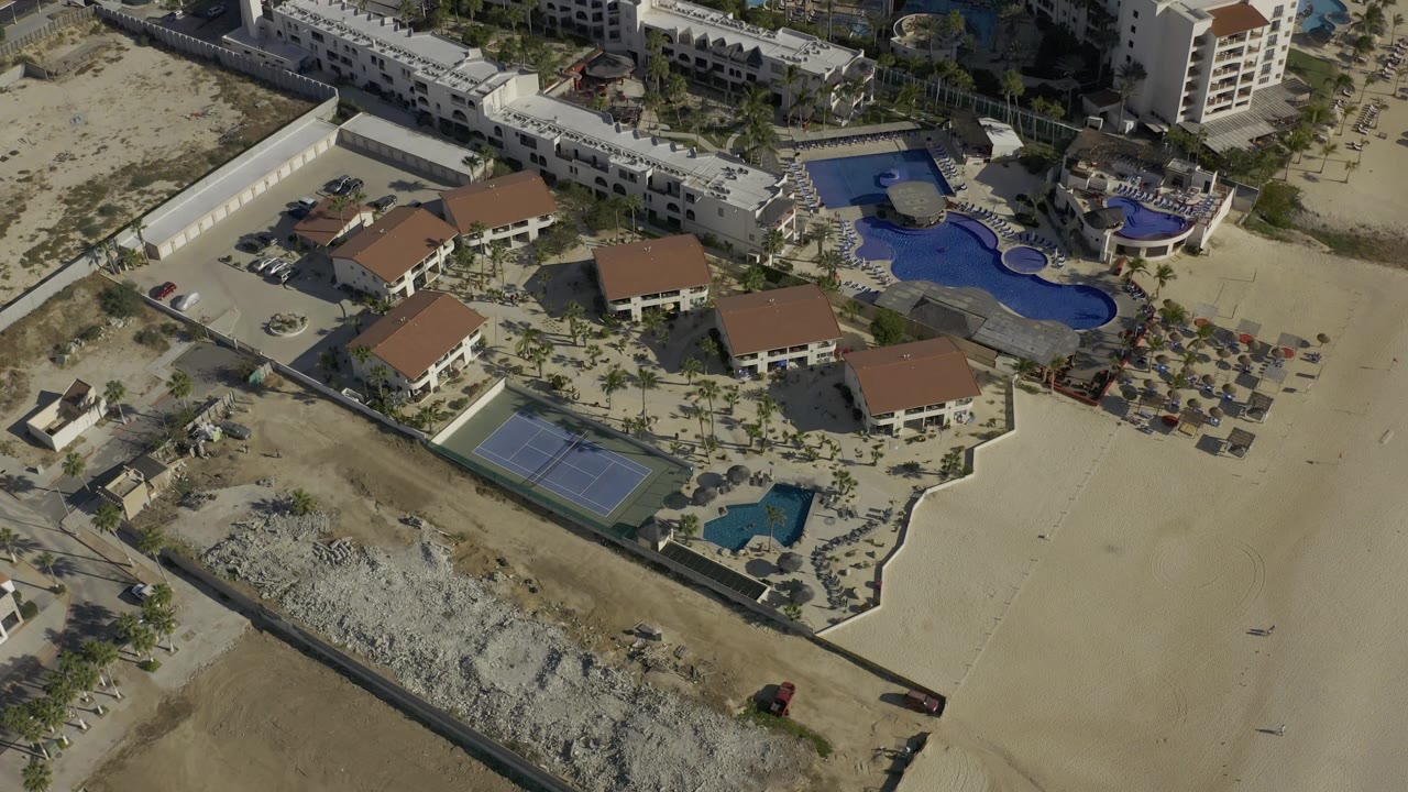 Aerial view in Baja California Sur, Mexico with the tennis court and small pool lower in frame, larger blue pools near white buildings, and no visible people.