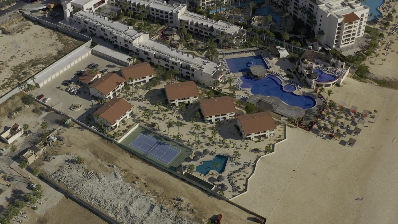 High oblique aerial frame in Baja California Sur, Mexico showing curved blue pools beside white buildings, red-roofed villas, sandy lots, and no visible people.