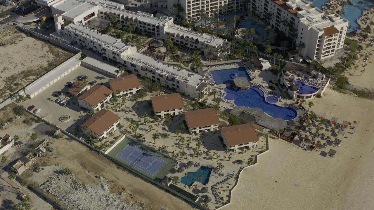 Oblique aerial view of buildings, pools, beach chairs, and sandy property edges.