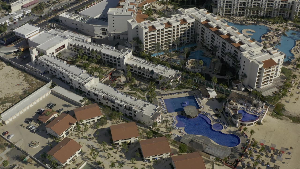 Aerial frame in Baja California Sur showing white resort buildings around curved blue pools, interior landscaping, sandy ground, and no visible people.