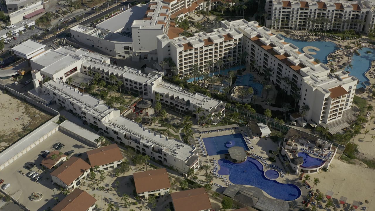 Top-view Baja California Sur resort frame with roof terraces, turquoise and dark blue pool shapes, perimeter roads, cars, and no visible people.
