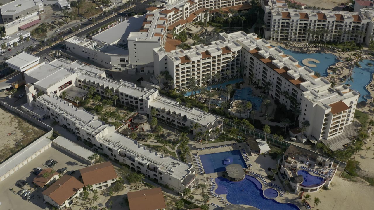 Overhead frame of a Baja California Sur hotel complex with a large blue pool deck, multi-story cream buildings, open sandy parcel, and no visible people.