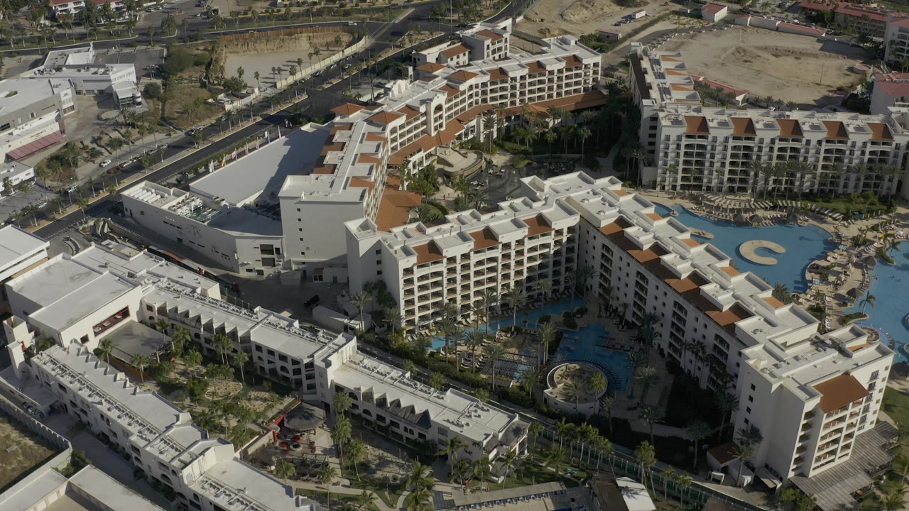 High oblique aerial frame in Baja California Sur, Mexico, showing white and tan resort buildings around bright blue pools, with roads and sandy lots beyond and no visible people.