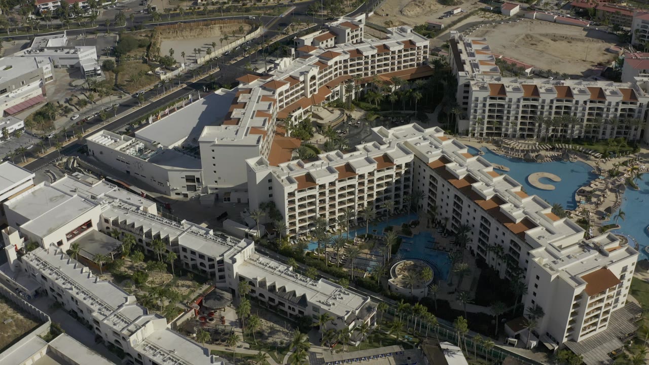 Aerial frame in Baja California Sur, Mexico, with the resort roofs and pool courtyards centered, a road corridor behind them, sandy parcels, and no visible people.