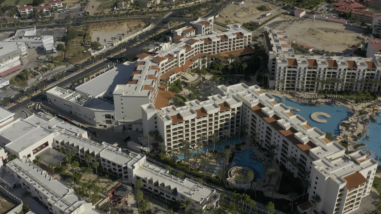 High aerial view in Baja California Sur, Mexico, showing the resort complex from a slightly shifted angle, with blue pools at right, white buildings across the foreground, and no visible people.