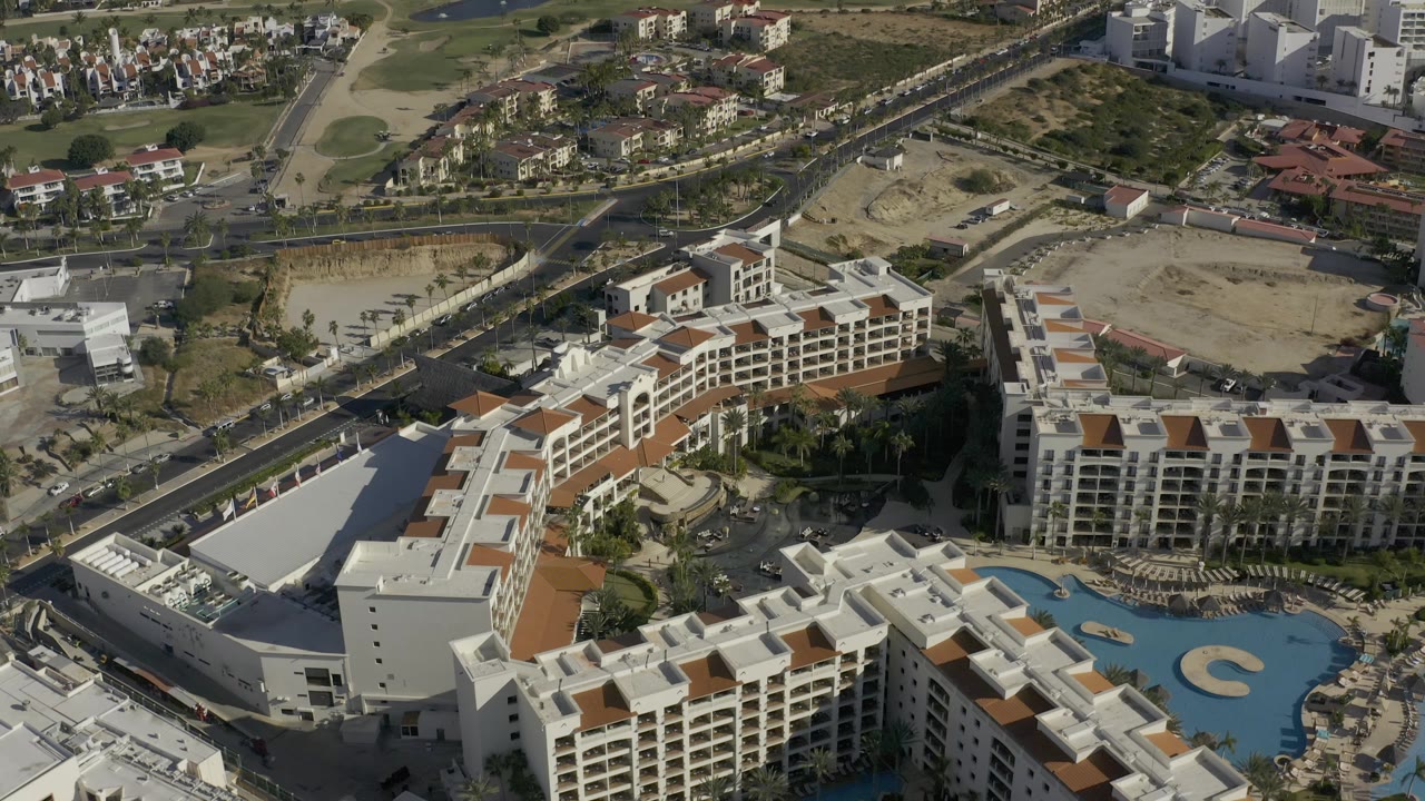 Second aerial frame in Baja California Sur with terracotta-roofed resort blocks, a blue pool, nearby roads, golf landscaping, dry construction-like lots, and no visible people.