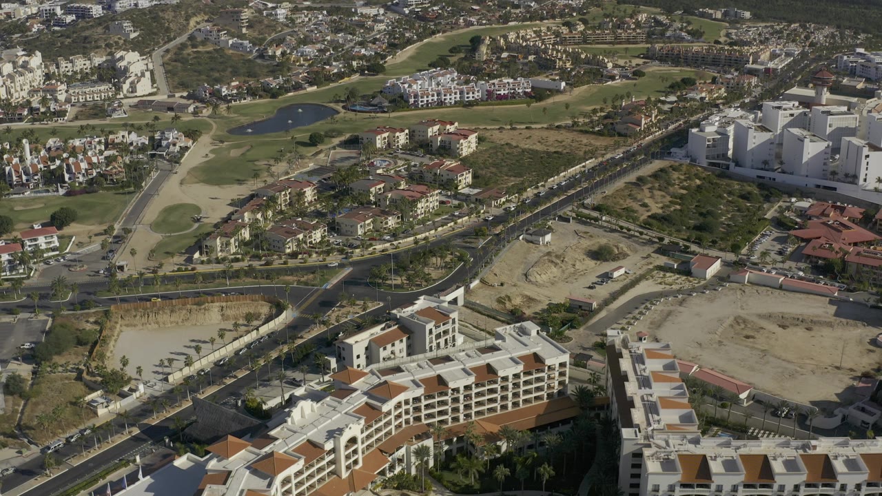 High overhead Baja California Sur view with bright white buildings at right, open dirt lots, golf greens and ponds at left, hillside homes in back, and no visible people.