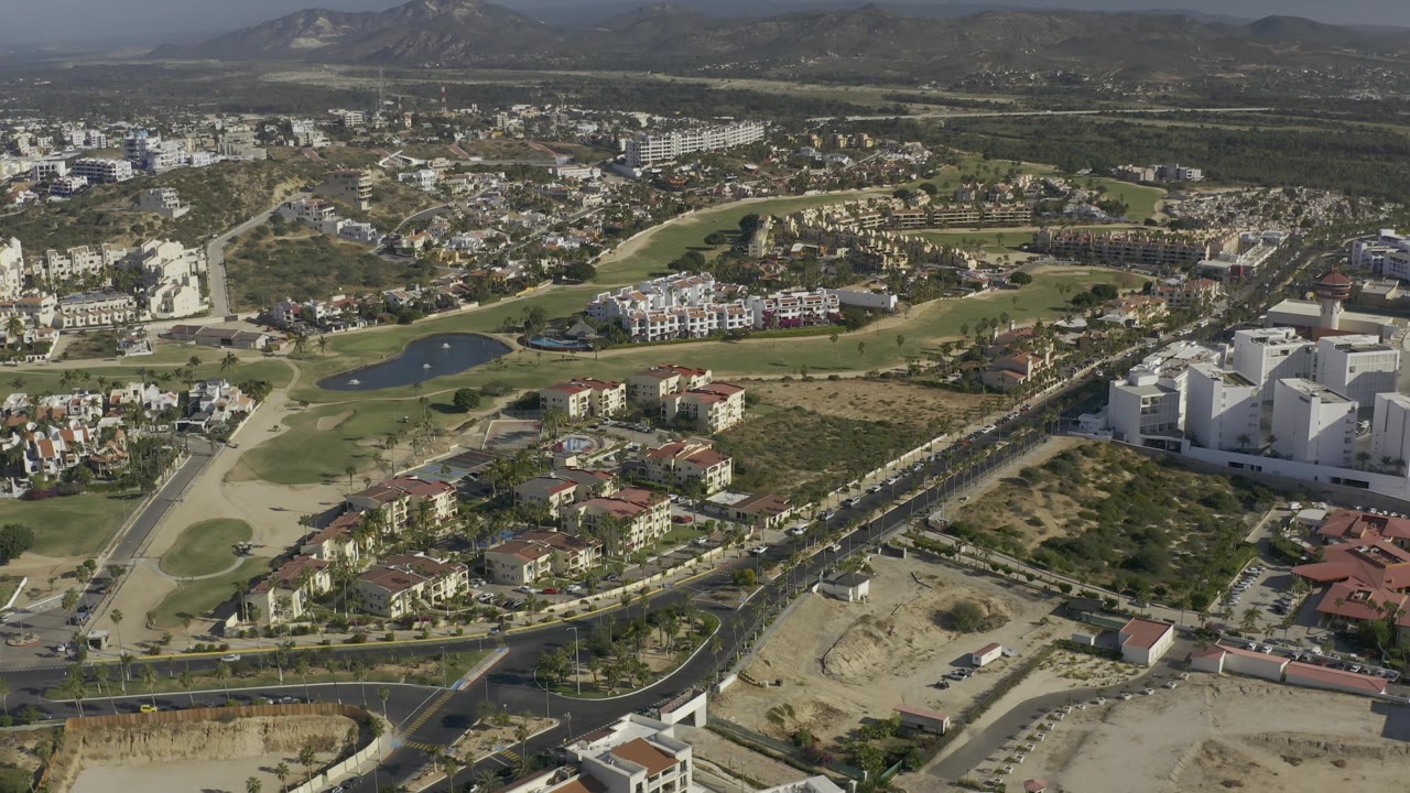 High drone-style view in Baja California Sur with golf-course greens and ponds centered among residential streets, dry undeveloped land in front, white buildings at right, and no visible people.