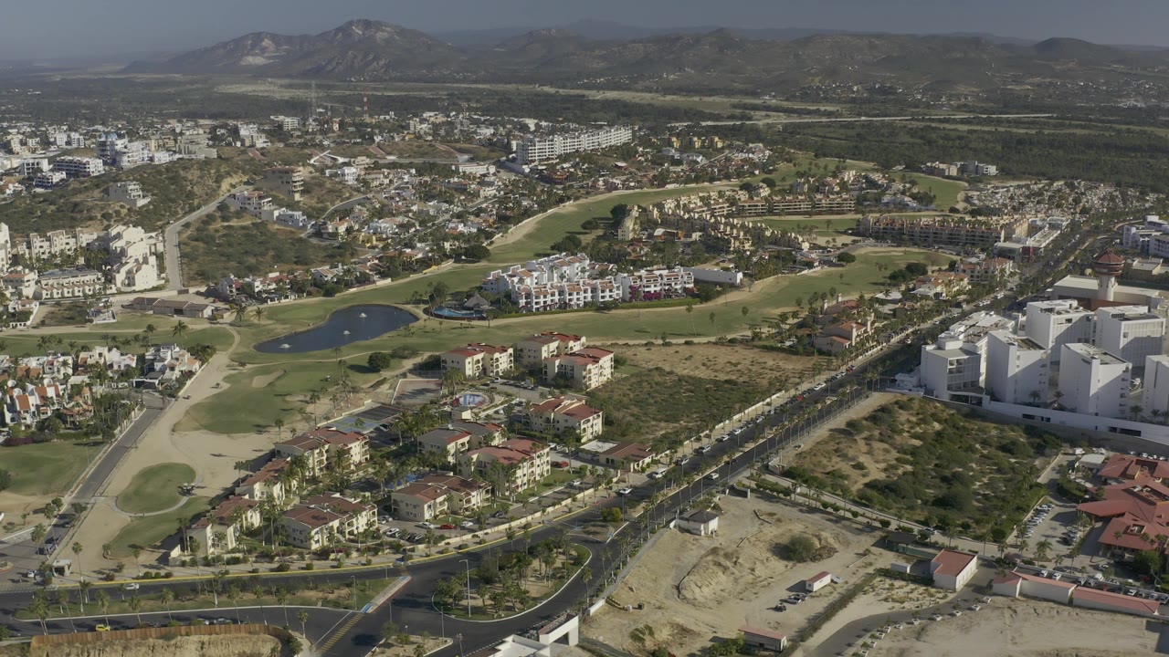 Wide aerial of a road junction, homes, golf greens, and low mountains.