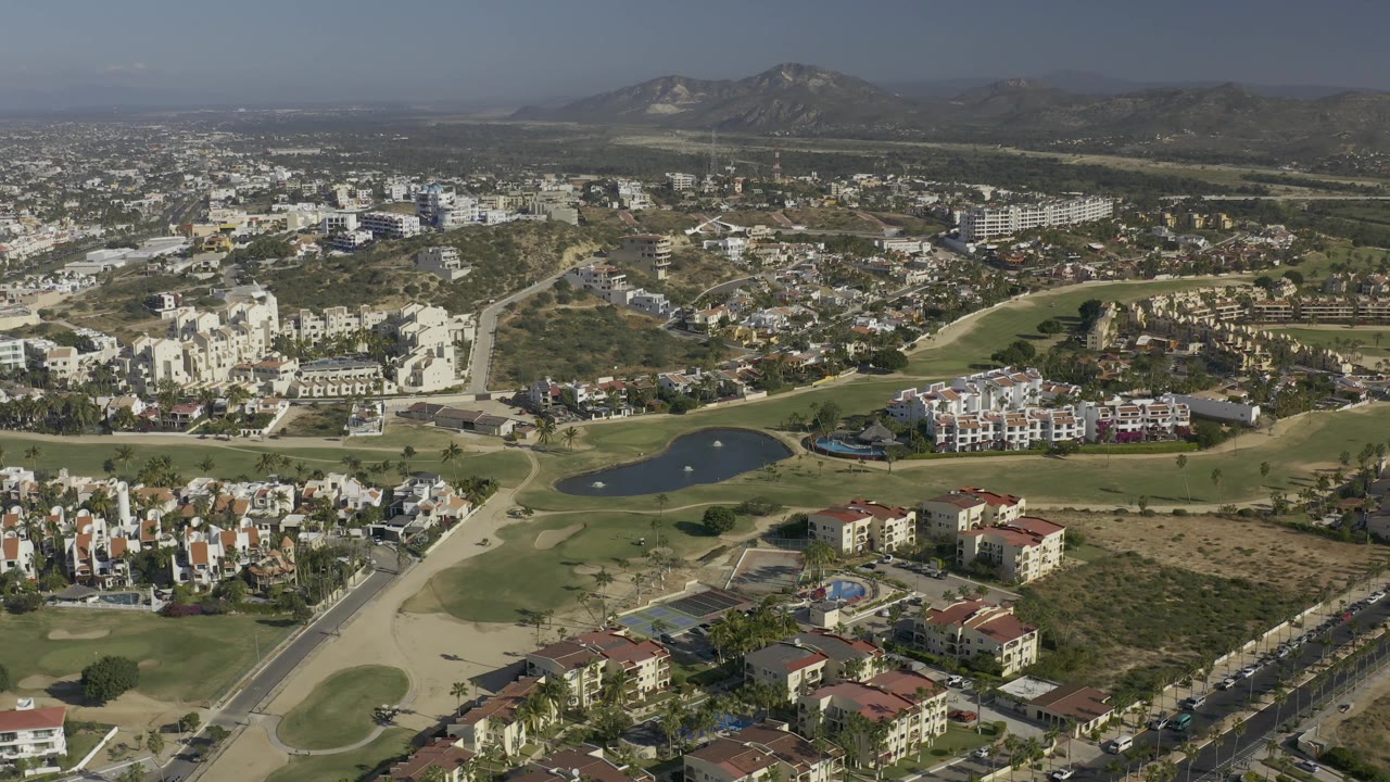Aerial frame from Baja California Sur, Mexico showing curved roads leading past a small lake, pale buildings, clustered homes, green fairways, dry terrain, hazy daylight, and no visible people.