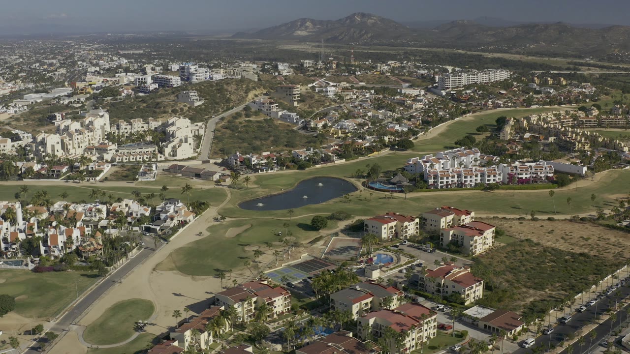 High aerial frame over Baja California Sur residential buildings beside golf fairways, a small pond, roads, dry lots, and hazy hills, with no visible people.