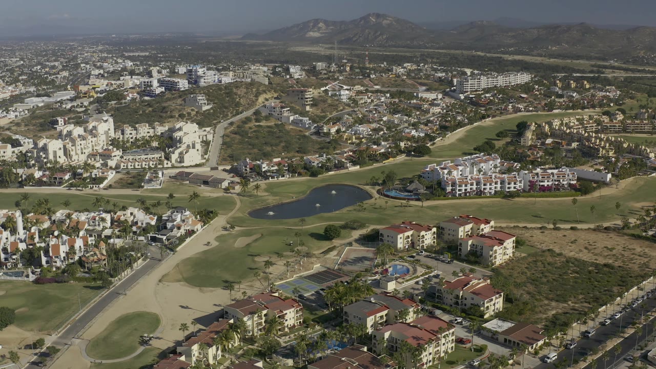 Drone-height view in Baja California Sur looking across pale apartment-style buildings, green fairways, a dark pond, roadways, dry ground, and distant hills, with no visible people.