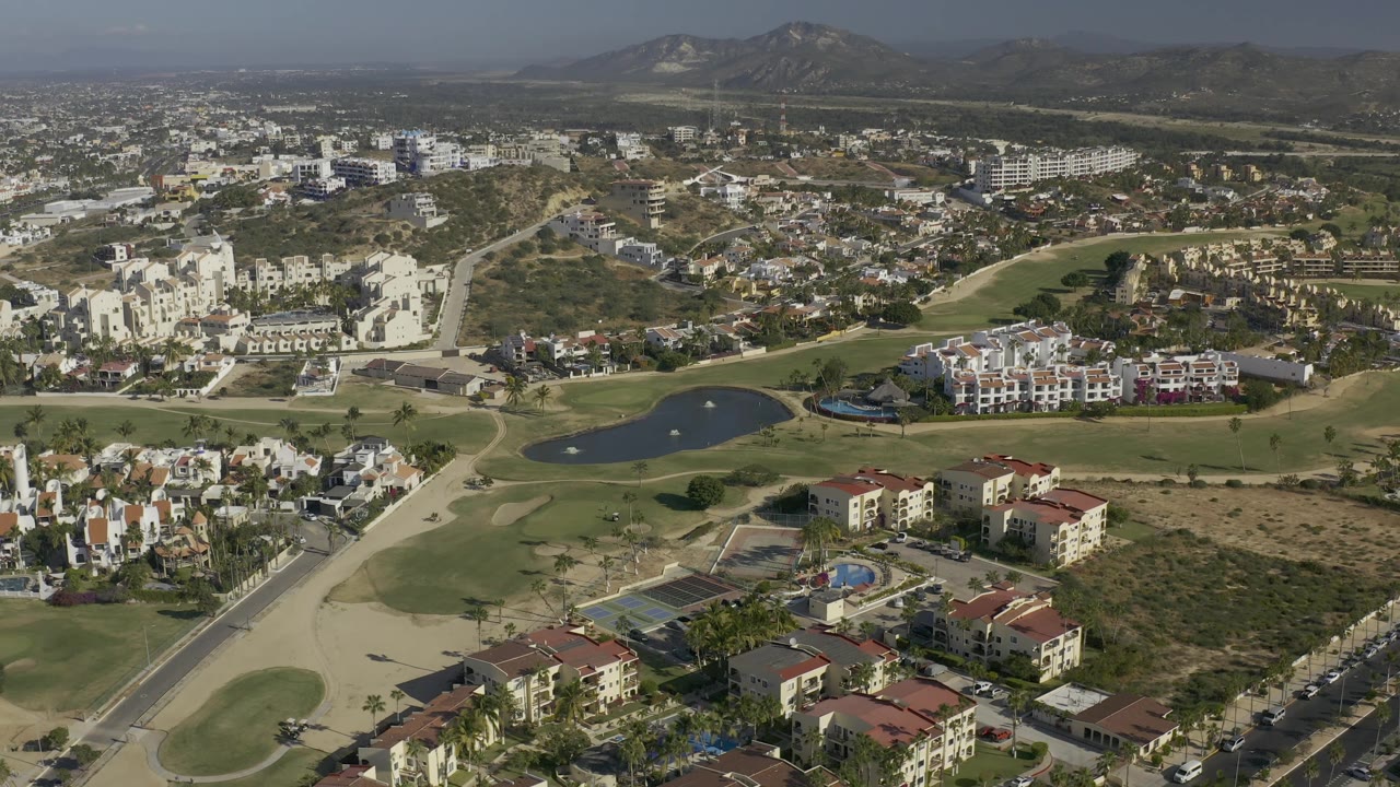 Wide aerial frame over Baja California Sur showing curving golf fairways, white residential blocks, villas, open dry parcels, roads, a pond, and a hazy mountain backdrop, with no visible people.