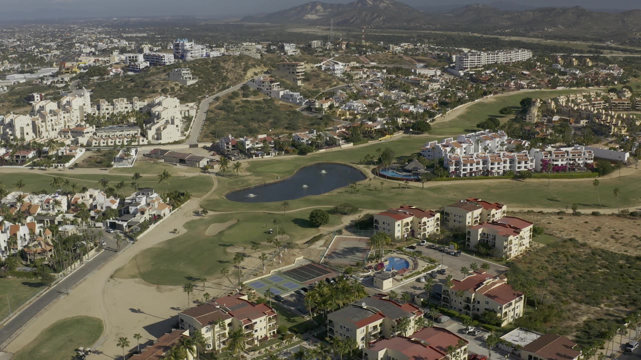 Aerial frame from Baja California Sur, Mexico showing red-roofed residences in the foreground, a dark golf-course lake in the midground, hillside buildings beyond, and no visible people.