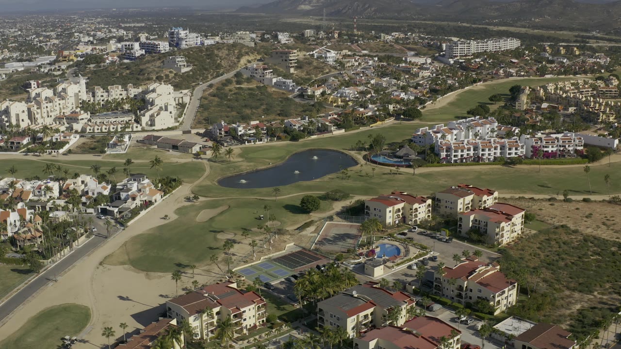Wide Baja California Sur aerial showing fairways and sand-colored cart paths around a dark lake, apartment-style buildings at the lower edge, dry lots to the right, and no visible people.