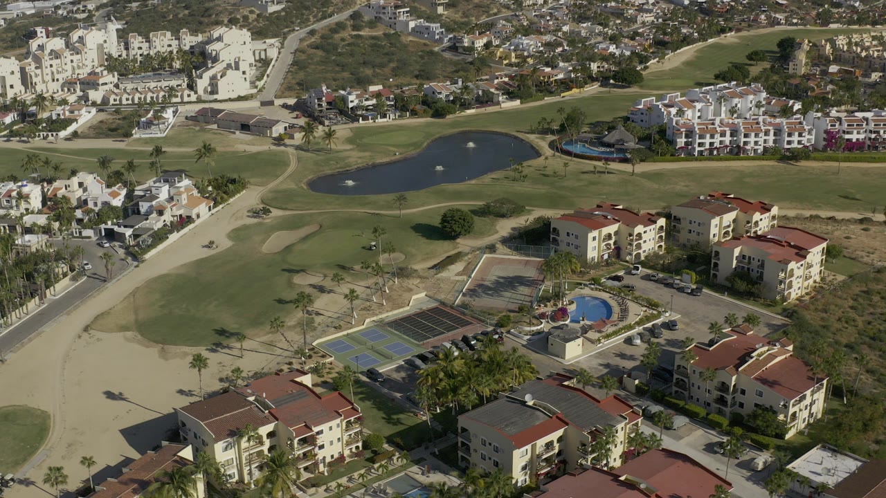 High aerial frame over Baja California Sur, Mexico, showing red-roof apartment buildings, tennis courts, a blue pool, golf fairways, ponds, roads, and surrounding homes.