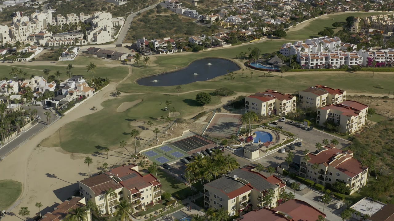Aerial frame in Baja California Sur, Mexico, with the apartment blocks lower in view, tennis courts and pool centered, and golf fairways, ponds, and residential streets beyond.