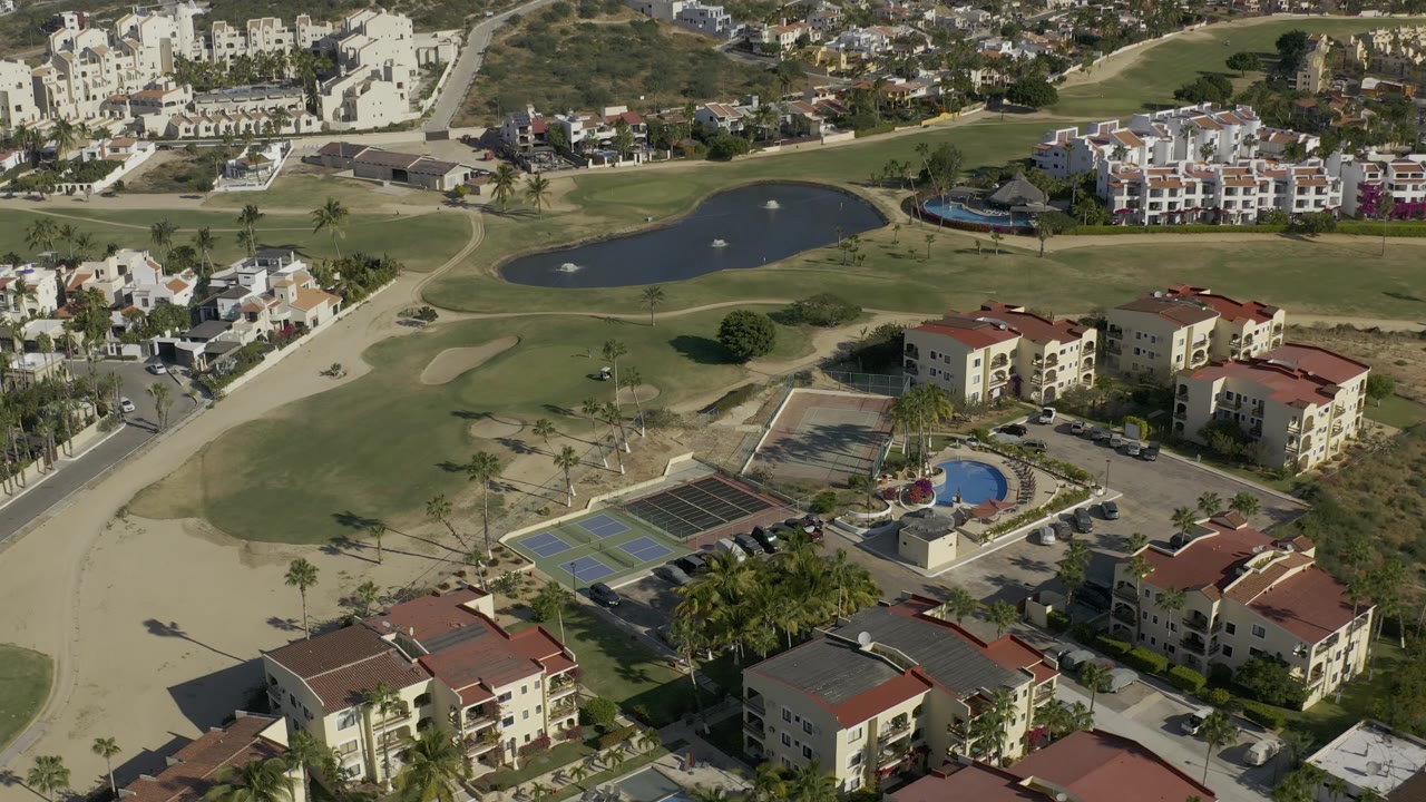 High overhead frame from Baja California Sur, Mexico, showing the apartment complex at the bottom, courts and pool in the middle, and fairways, ponds, and clustered homes in the background.