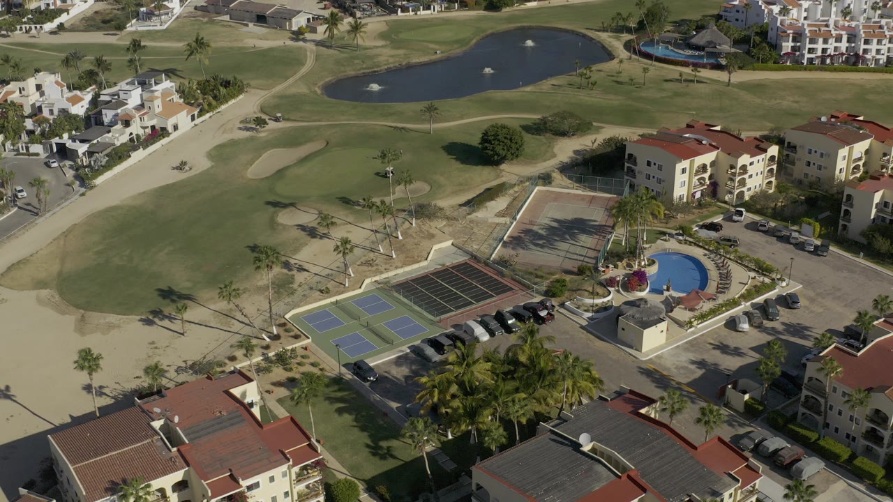 High aerial frame over a Baja California Sur golf course area with tennis courts, a round blue pool, parked cars, palm trees, red-roof buildings, and no visible people.