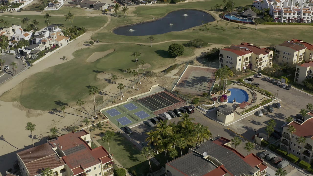 Drone view of fairway, pond, tennis courts, pool, roads, and low-rise buildings.