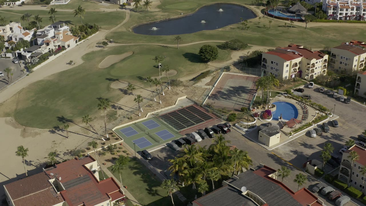 Baja California Sur aerial frame showing the golf course pond and sand traps above a recreation area with courts, a pool, palm trees, parked vehicles, and no visible people.