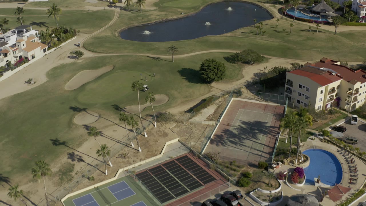 High aerial frame over a Baja California Sur golf course with tennis courts in the foreground, fairways and bunkers beyond, a dark pond near the top, and no visible people.