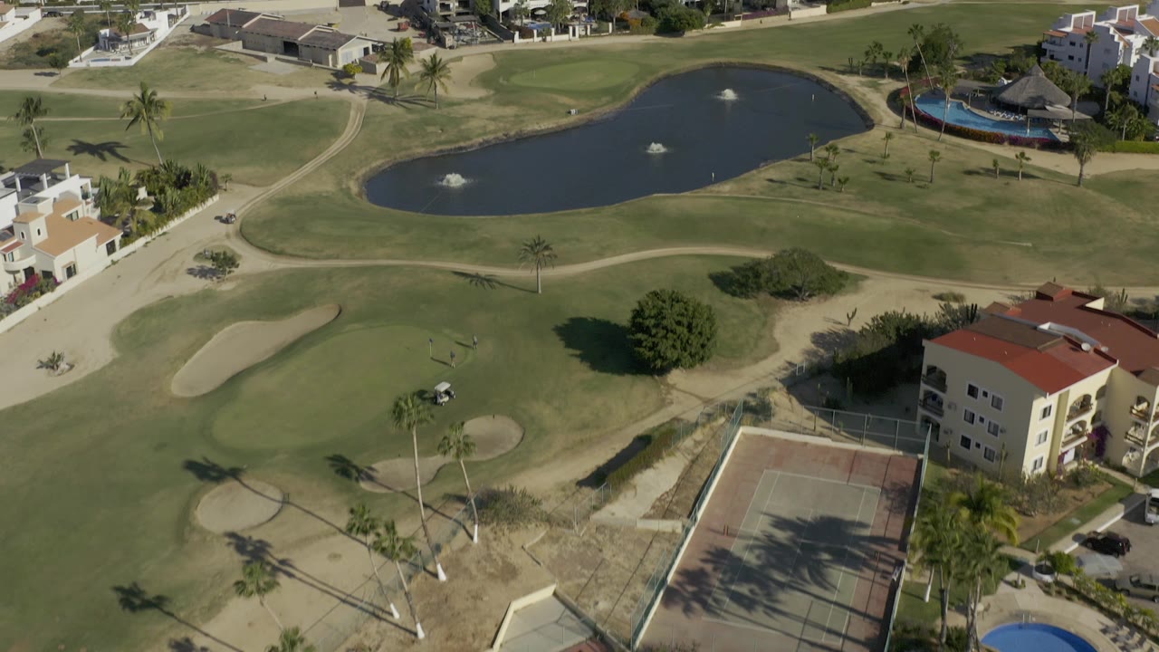 High drone view of the Baja California Sur course with a broad fairway and bunkers in the center, a pond at the top, residential buildings on the right, courts at the bottom, and no visible people.