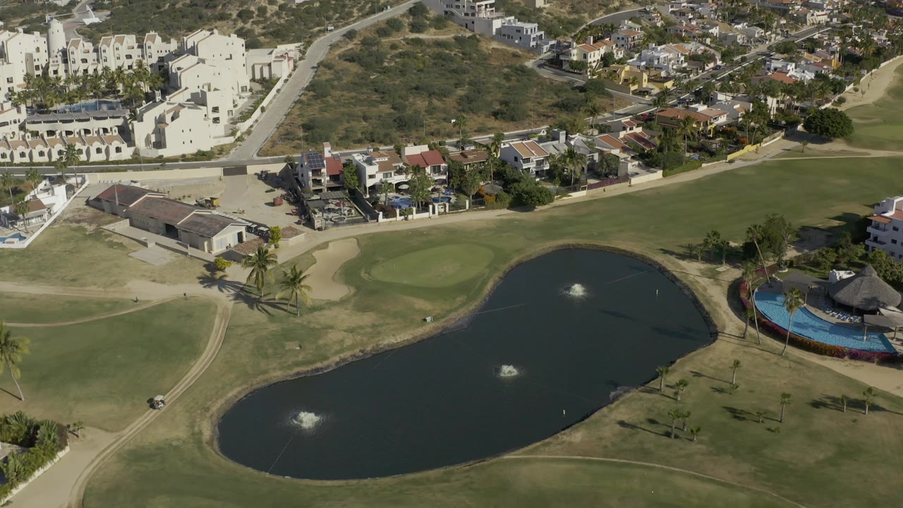 Aerial frame in Baja California Sur, Mexico, with the lake centered lower in view, two fountain sprays visible, fairway grass around it, hillside homes beyond, and no visible people.