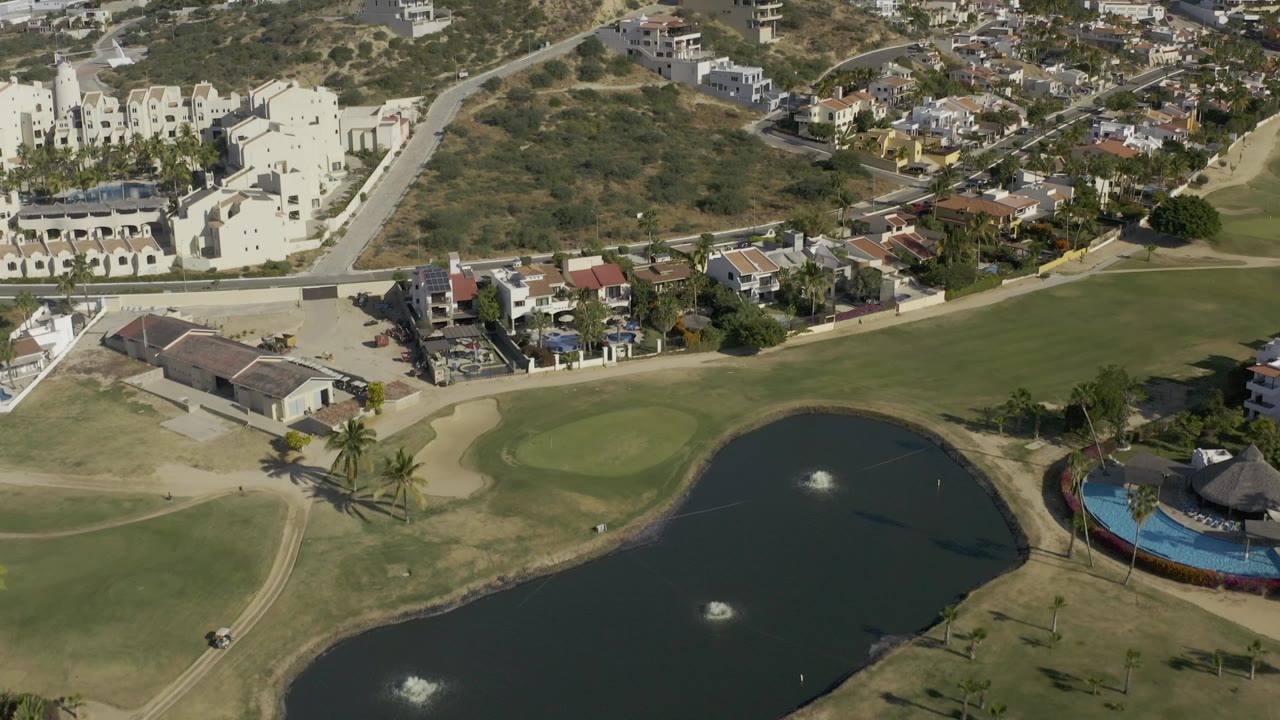 Wide aerial view in Baja California Sur, Mexico, with the lake shifted to the right, a putting green and palm trees on the left, hillside residences above, and no visible people.
