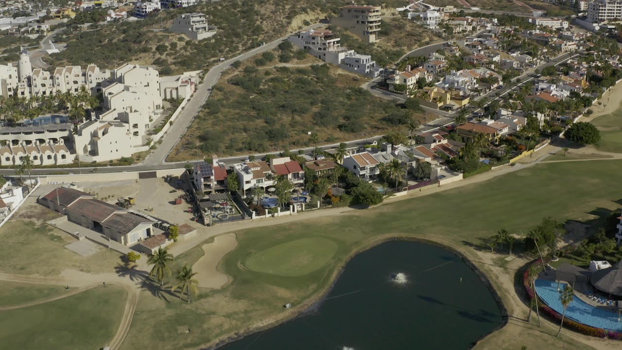 Aerial angle over white buildings, a green fairway, pond edge, and hillside neighborhood.
