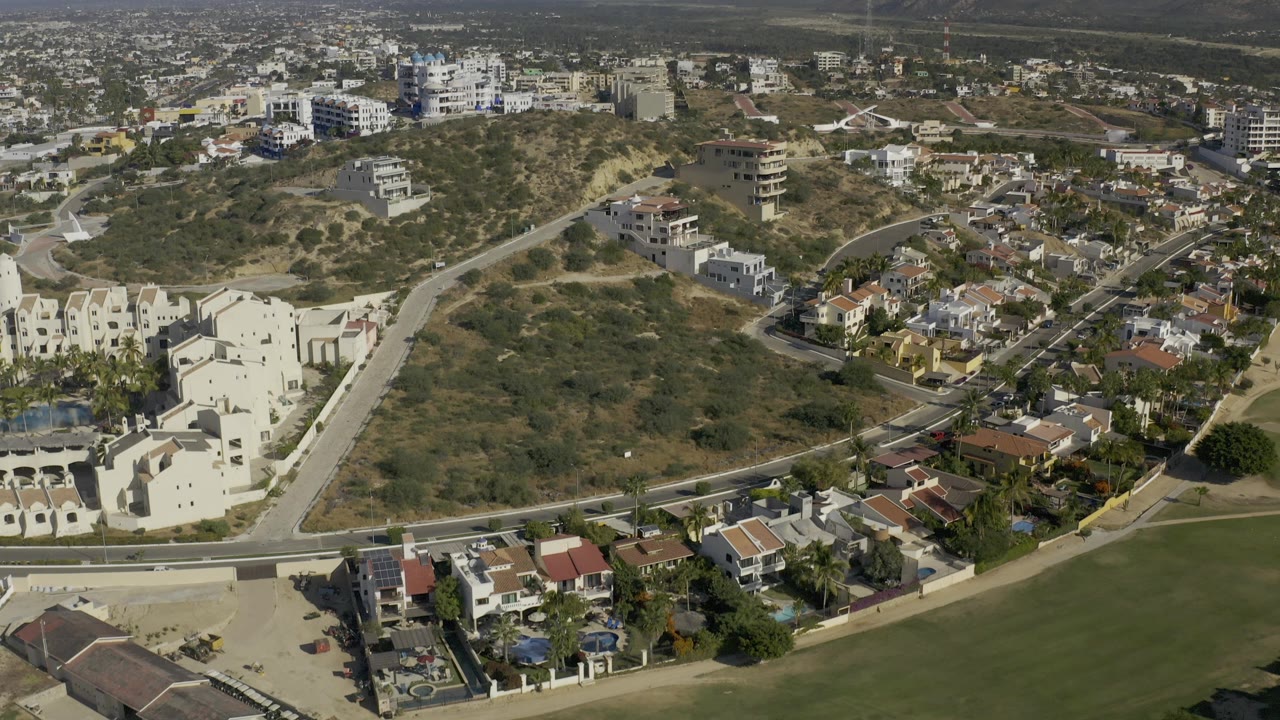 Aerial frame over Baja California Sur with cream apartment buildings, dry scrub hills, curving roads, distant town blocks, hazy mountains, and no visible people.