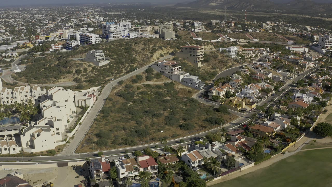 Aerial view of Baja California Sur with a scrub-covered hill centered between low homes, larger white buildings, a hazy urban background, and no visible people.