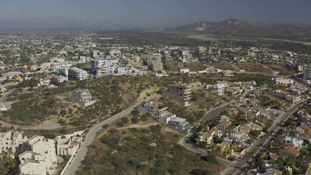 Aerial view in Baja California Sur with hillside homes lower in frame, a winding road through dry brush, denser town blocks beyond, mountains on the horizon, and no visible people.
