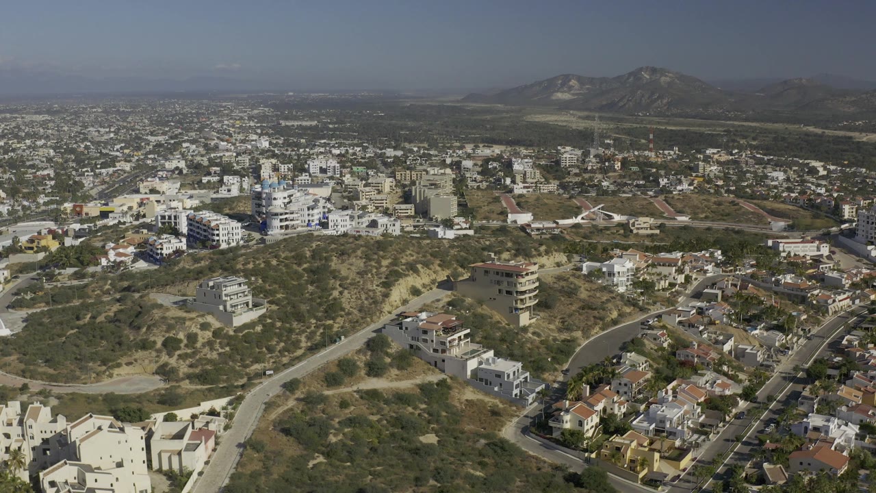 Mid-clip aerial frame in Baja California Sur showing low-rise white and tan buildings, scrubby terrain, paved roads, a broad settlement, distant mountains, and no visible people.