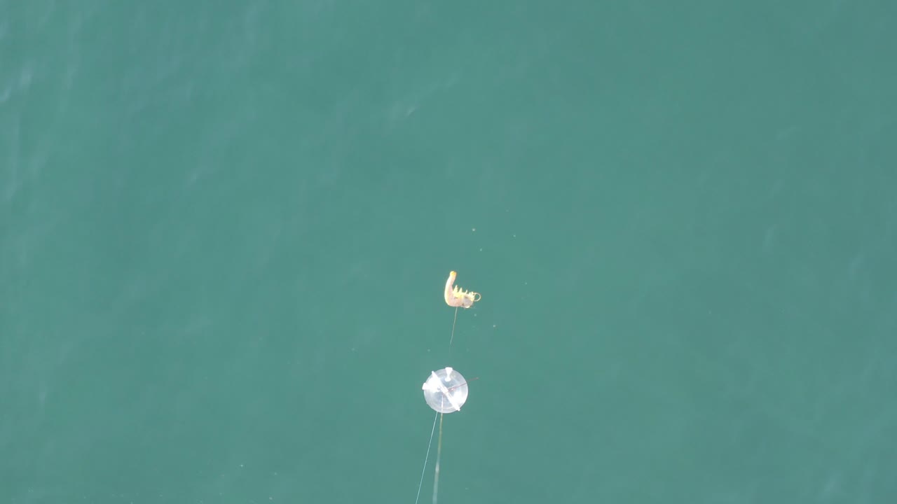 Overhead frame from Baja California Sur, Mexico, showing turquoise water, a white circular float, a small yellow bait float above it, and a person visible as a small shape near the gear.