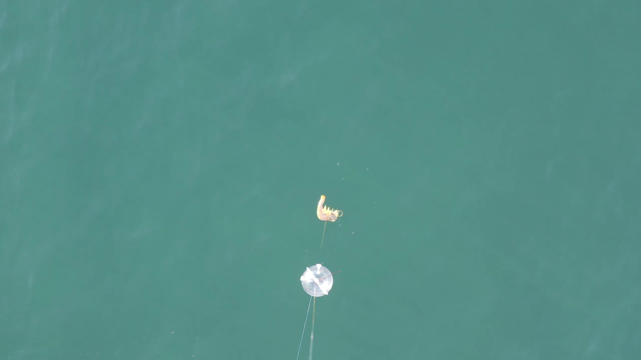 Aerial Baja California Sur frame with the white float near center, the yellow bait float above it, faint trailing lines, and the person still small against the blue-green water.