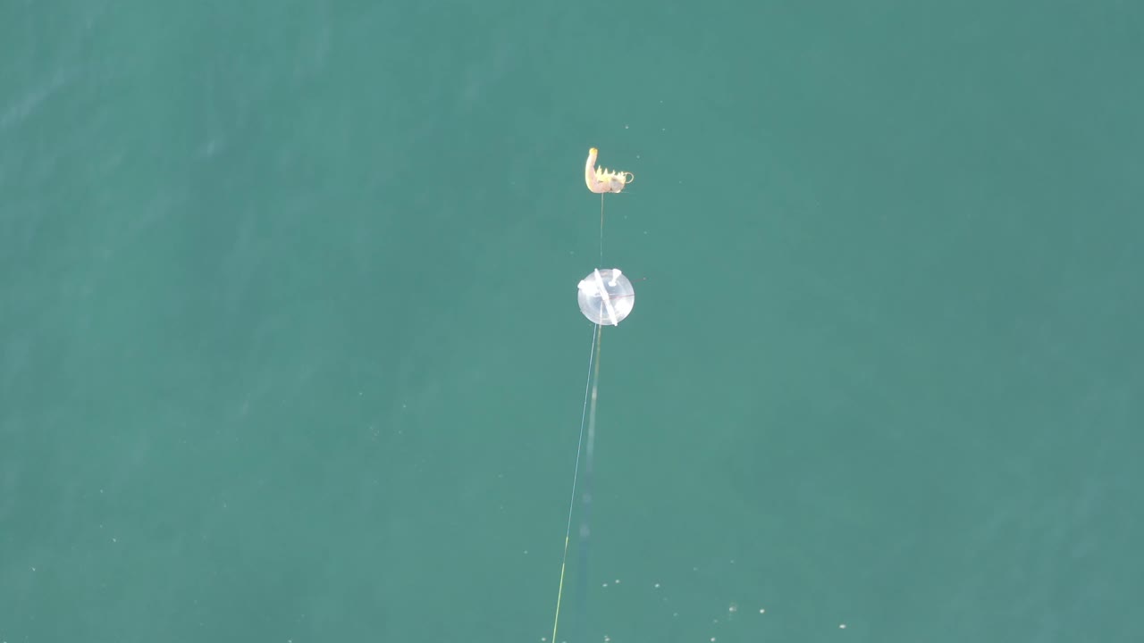 Aerial frame from Baja California Sur, Mexico, with the white circular object and pale curved object near the upper center, their lines extending downward through calm teal water and no visible people.