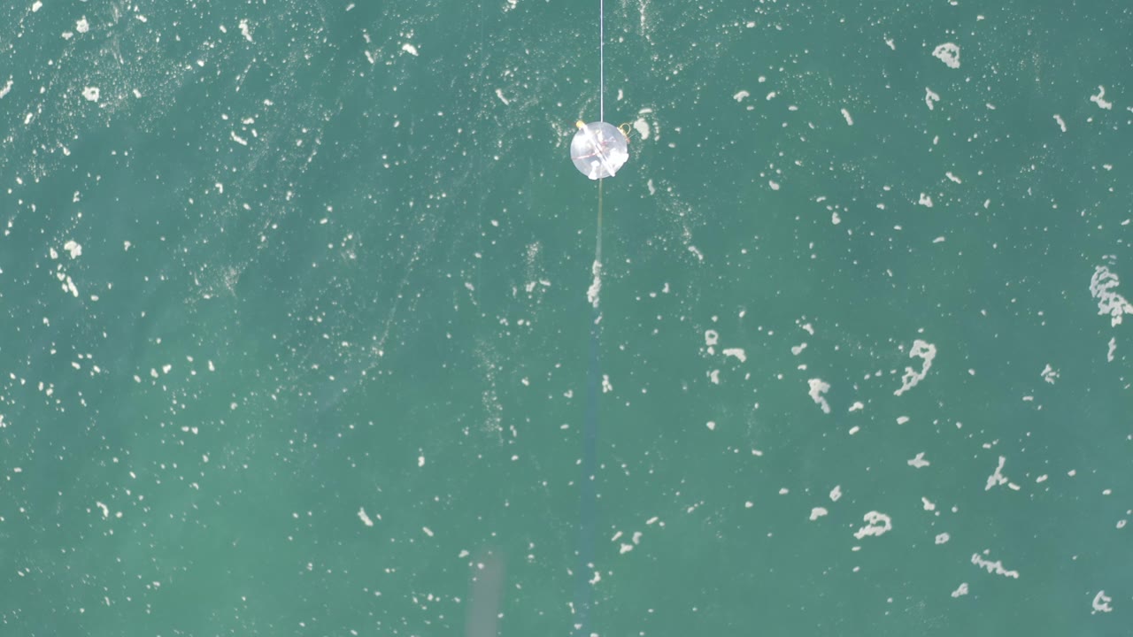 Overhead frame from Baja California Sur, Mexico, showing teal water, white foam streaks, and a cotton ball on a fine thread near the upper-right area with no visible people.