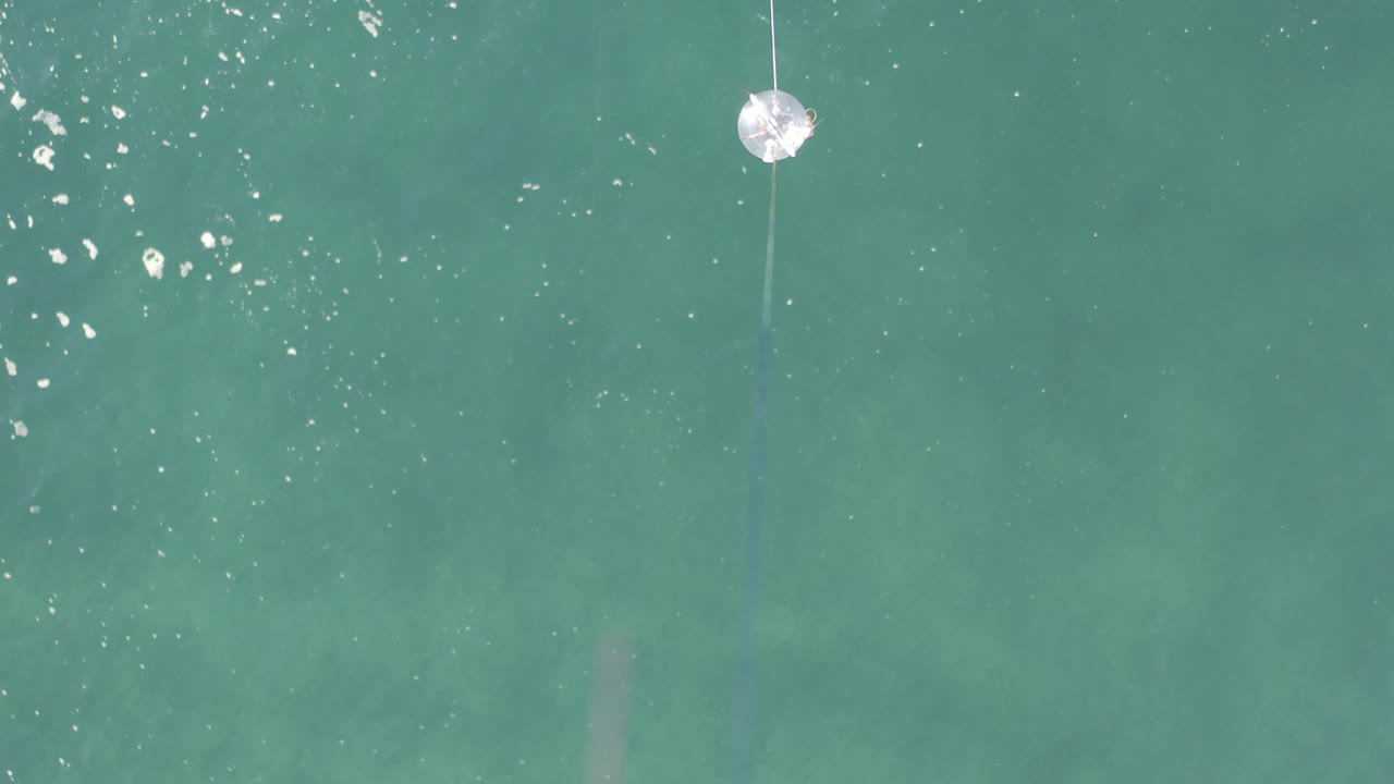 Overhead view of turquoise water in Baja California Sur, Mexico, with a small white circular floating object near the upper center, surface sparkle, faint underwater patches, and no visible people.