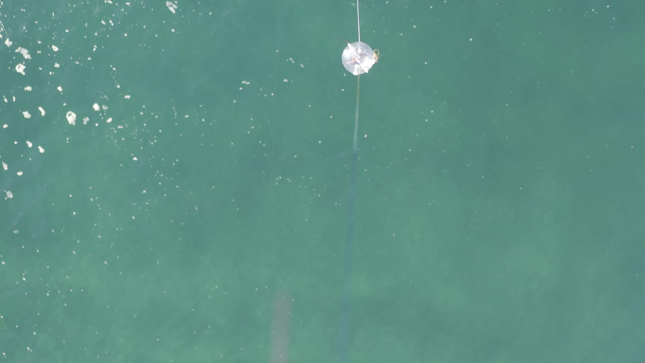 Aerial frame over blue-green water in Baja California Sur, Mexico, showing the white floating object near the top edge, a thin line dropping through the frame, scattered light flecks, and no visible people.