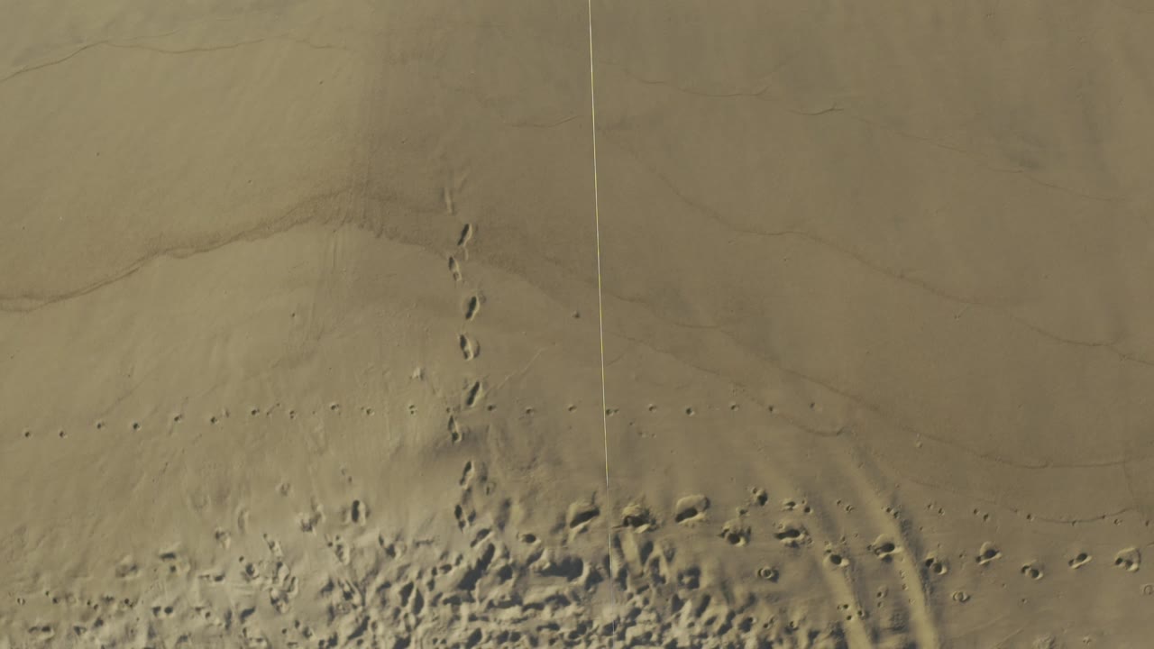 Overhead frame in Baja California Sur, Mexico, showing dense footprints across lower sand, broad curved tire-like tracks on the right, faint foam at the upper edge, and no visible people.