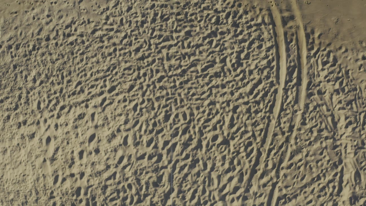 Top-down frame of beige sand in Baja California Sur, Mexico, showing dense footprints, compressed tire marks near the right edge, and no visible people.