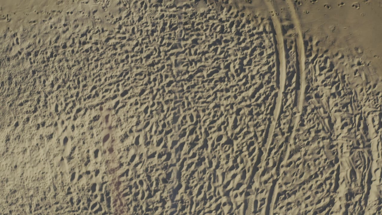 Aerial view of sandy ground in Baja California Sur, Mexico with a broad field of footprints, parallel track grooves on the right, and no visible people.