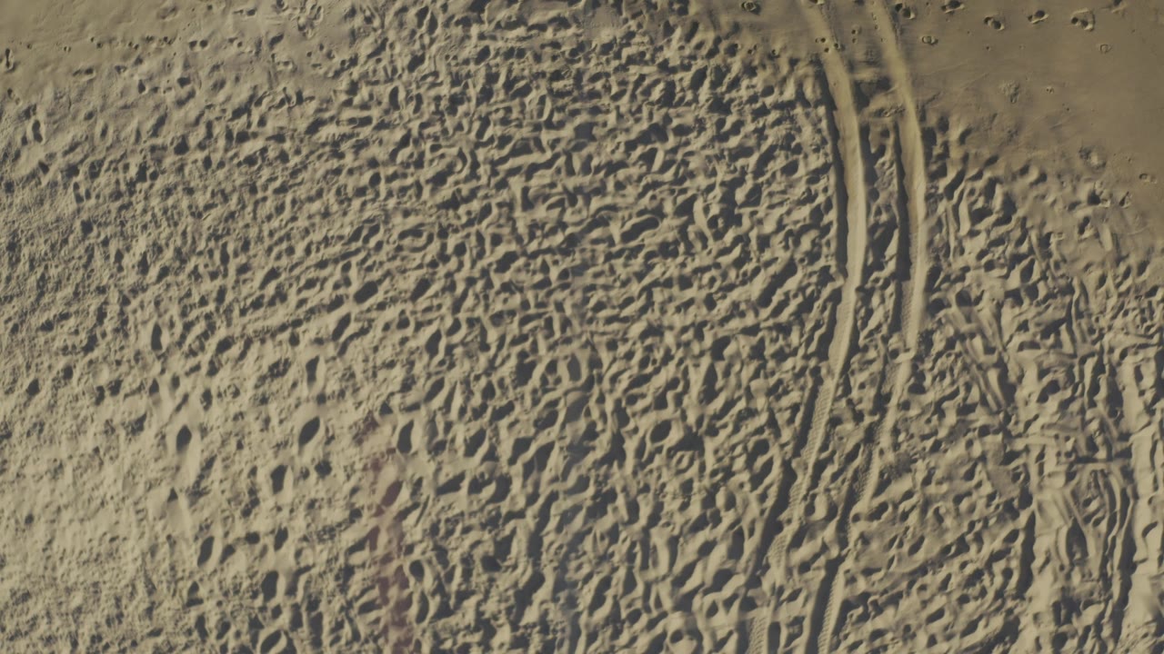 Sunlit sandy terrain in Baja California Sur, Mexico seen from above, with a curved tire track cutting through the right side and no visible people.