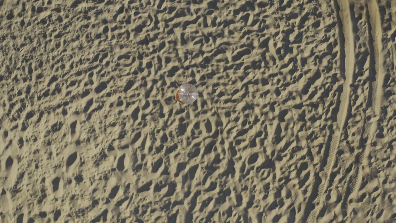 Aerial Baja California Sur desert view with the small pale object closer to the upper center, dense footprints or wind marks in the sand, tire tracks to the right, and no visible people.
