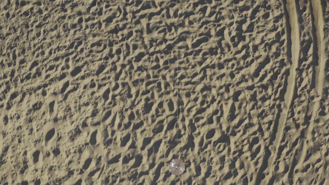 Top-down Baja California Sur desert frame with a faint pale object near the right side, parallel tire tracks along the edge, mottled sand texture, and no visible people.
