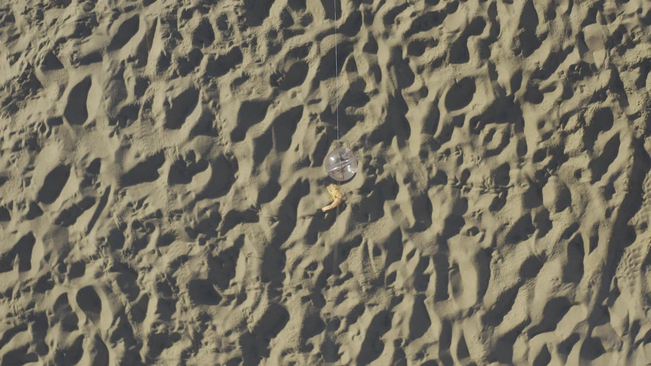 Ripples and footprints in pale beach sand with a small clear ball near the upper center.